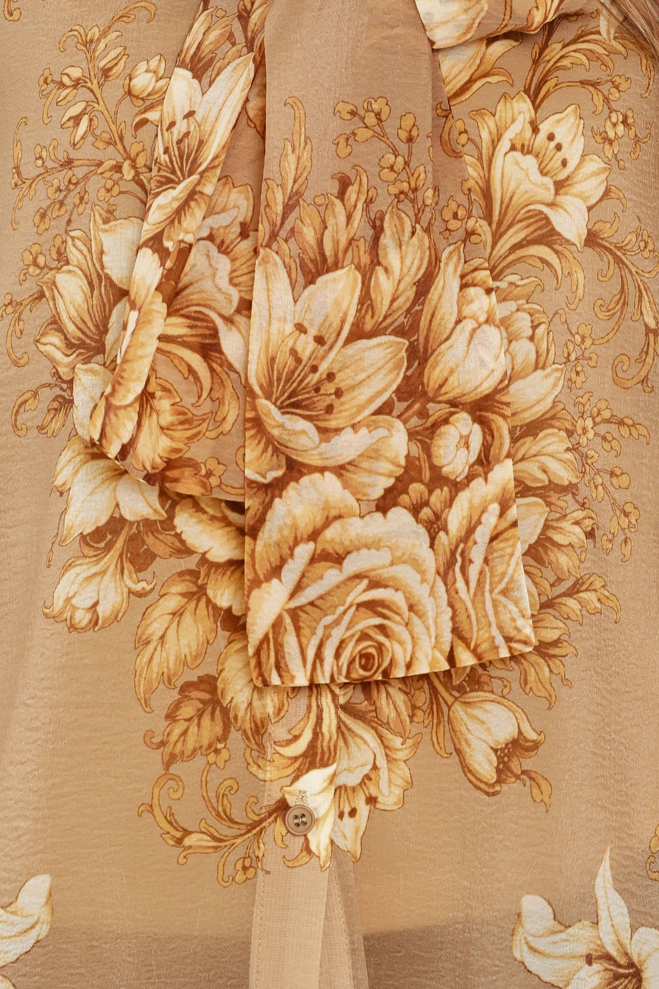 A woman wearing a sheer beige blouse with a large gold floral print and a bow tie neckline | Flying Tomato