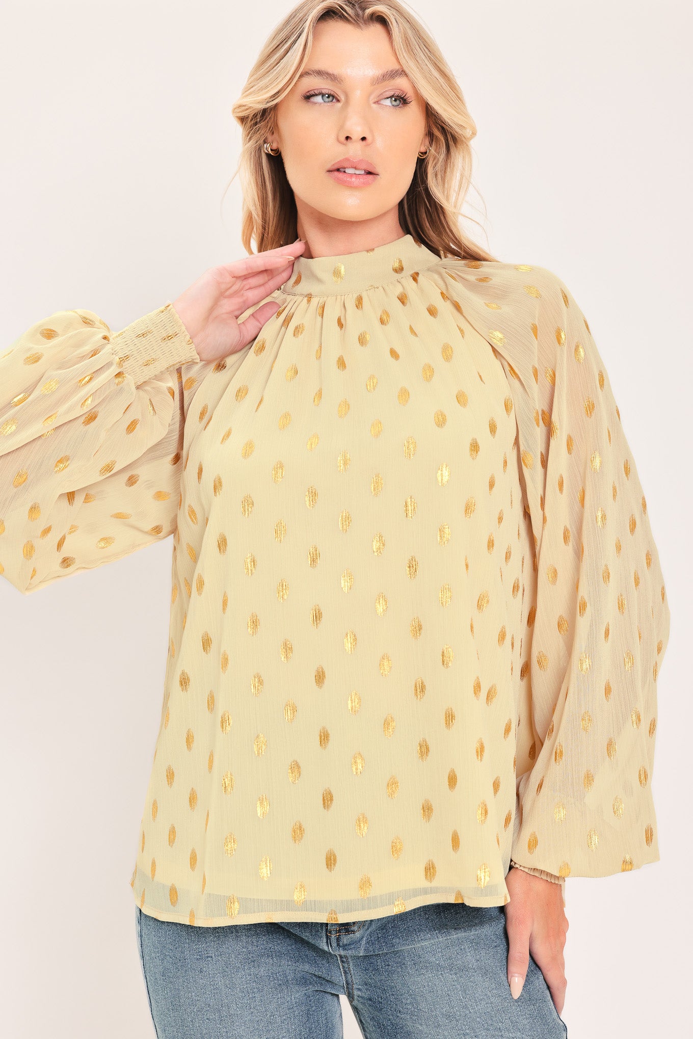 A woman wearing a light beige high-neck blouse with metallic gold polka dots and voluminous long sleeves | Flying Tomato