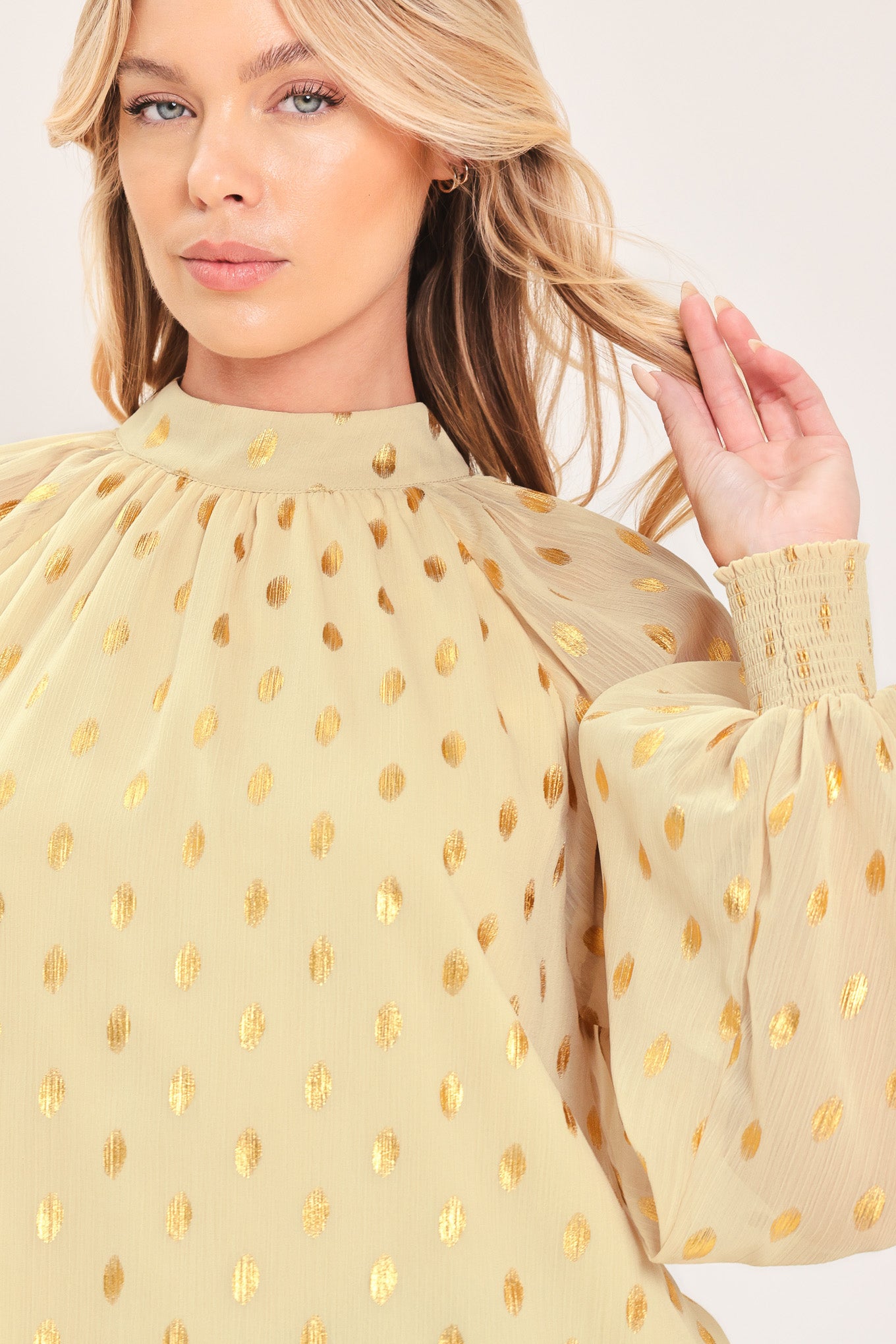 A woman wearing a light beige high-neck blouse with metallic gold polka dots and voluminous long sleeves | Flying Tomato