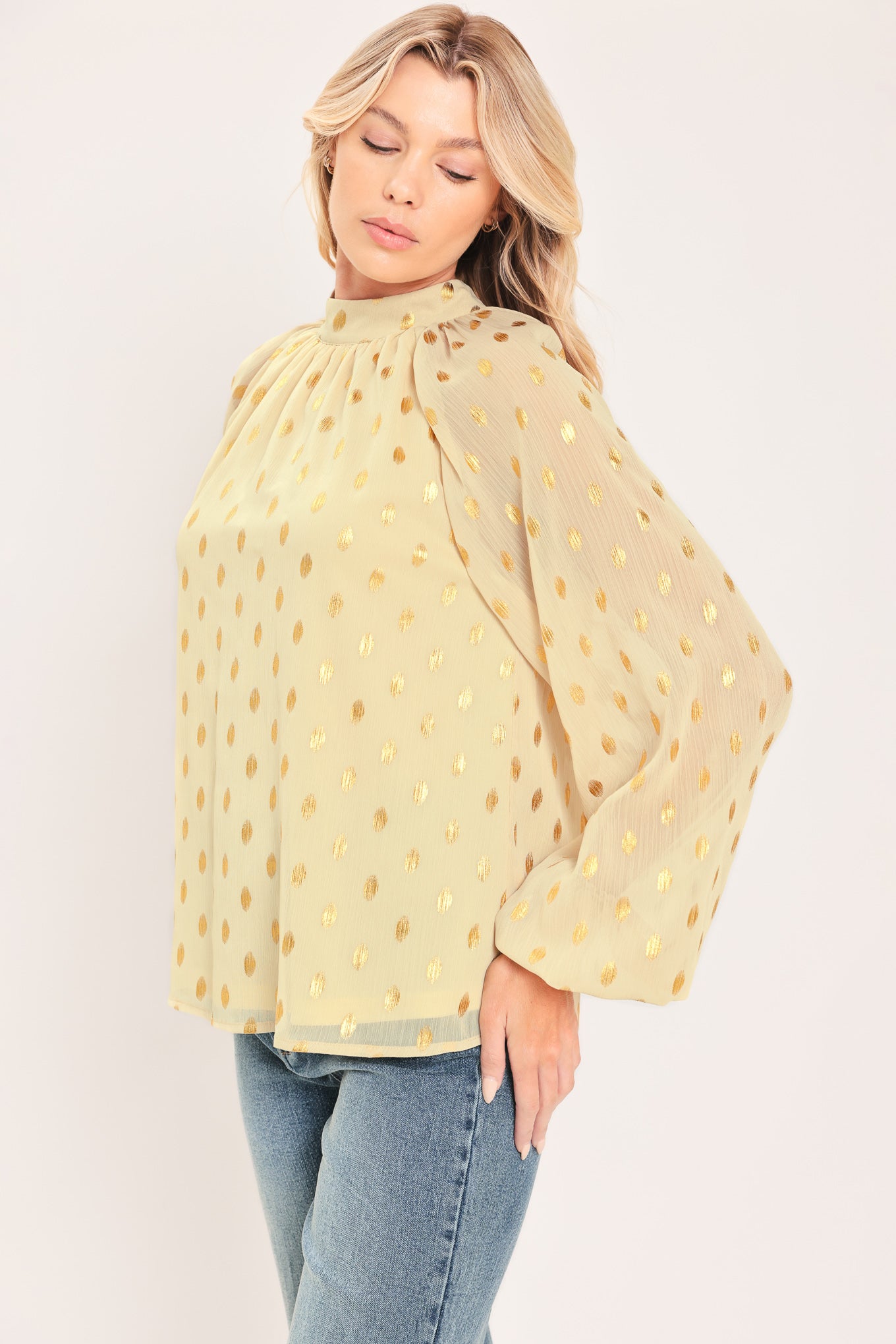 A woman wearing a light beige high-neck blouse with metallic gold polka dots and voluminous long sleeves | Flying Tomato
