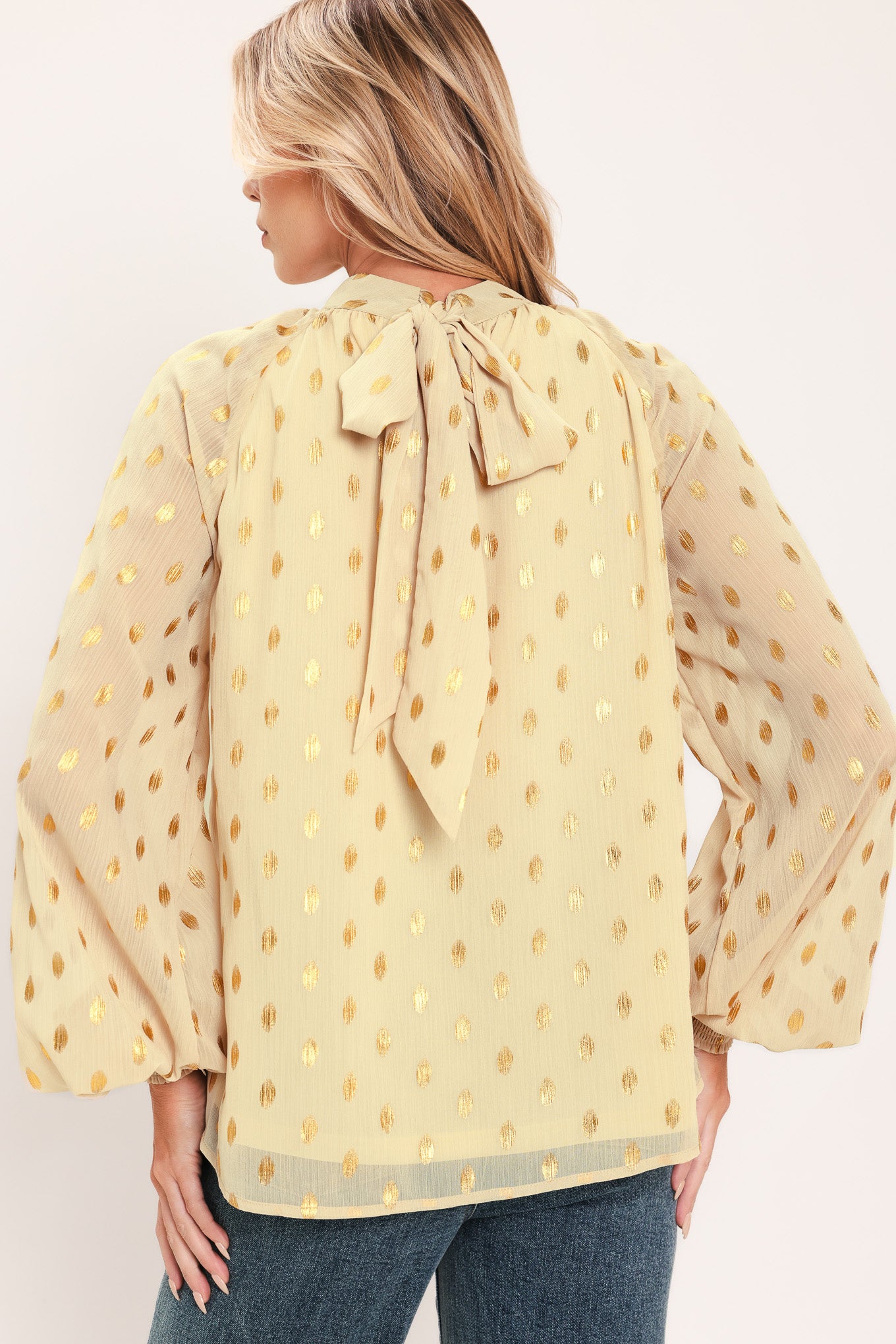 A woman wearing a light beige high-neck blouse with metallic gold polka dots and voluminous long sleeves | Flying Tomato