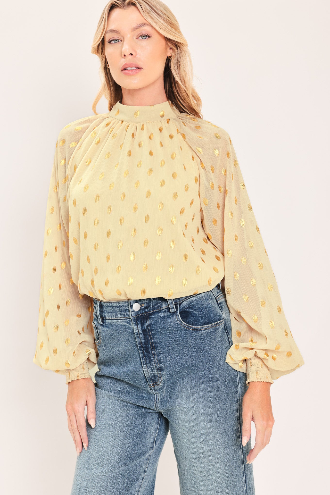 A woman wearing a light beige high-neck blouse with metallic gold polka dots and voluminous long sleeves | Flying Tomato