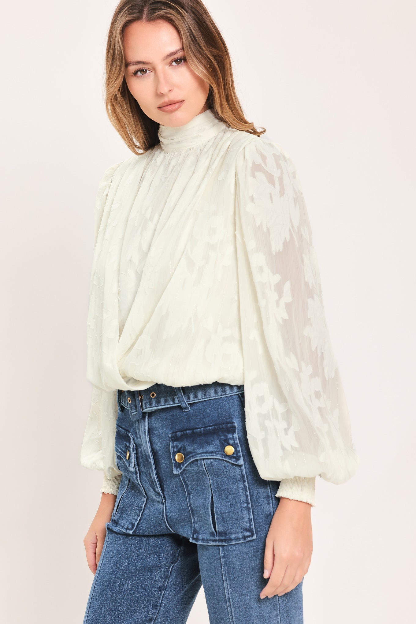 A woman wearing a cream-colored, semi-sheer long-sleeve blouse with a high neck | Flying Tomato