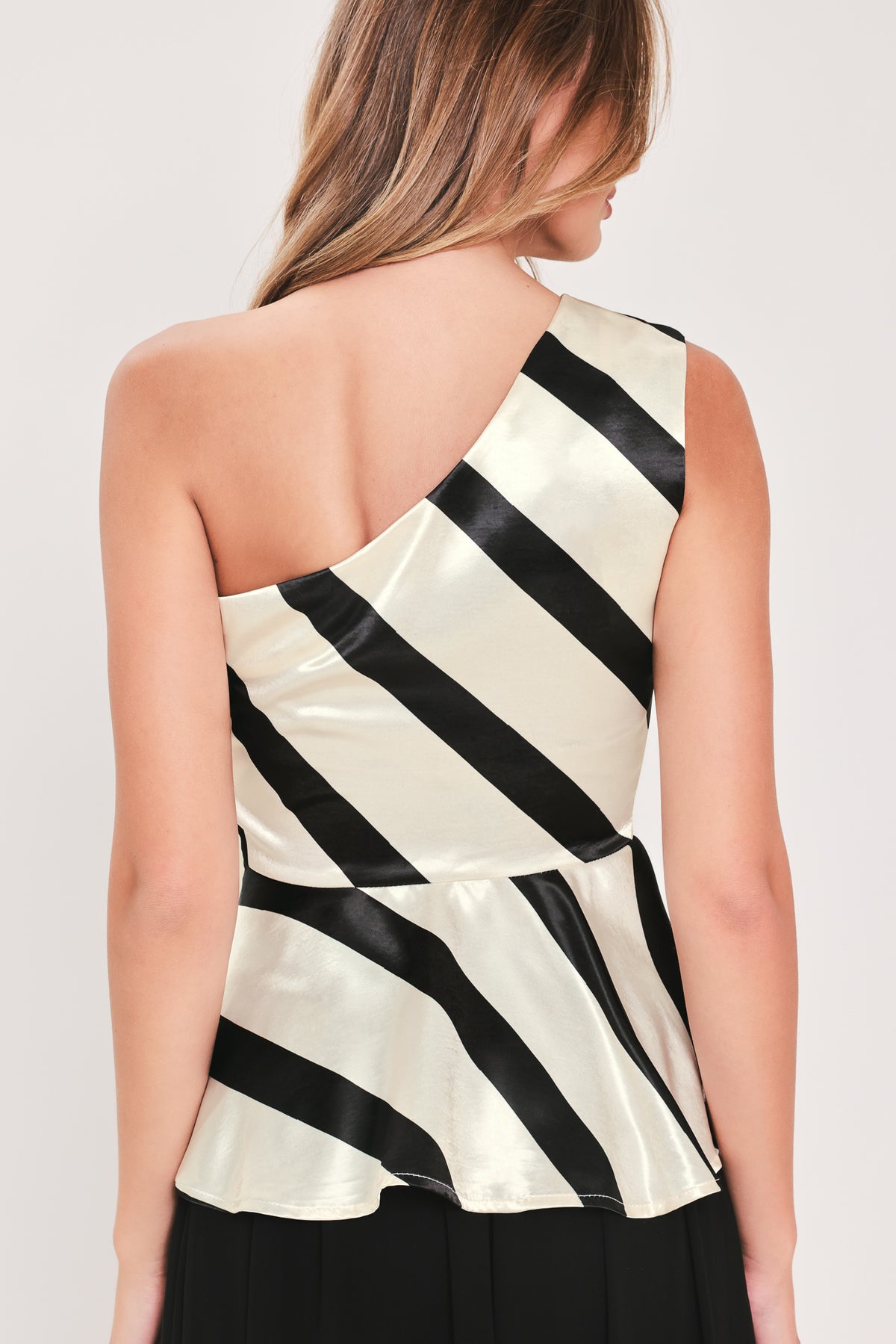 A woman modeling a sleeveless one-shoulder peplum top with diagonal black and cream satin-like stripes | Flying Tomato