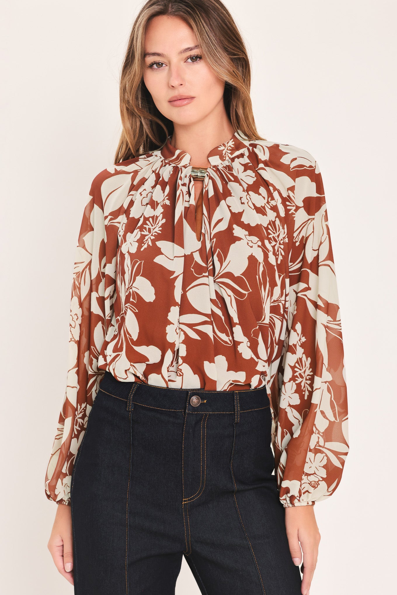 A woman wearing a rust-brown and cream floral print blouse with long sheer sleeves and a tie-neck detail | Flying Tomato