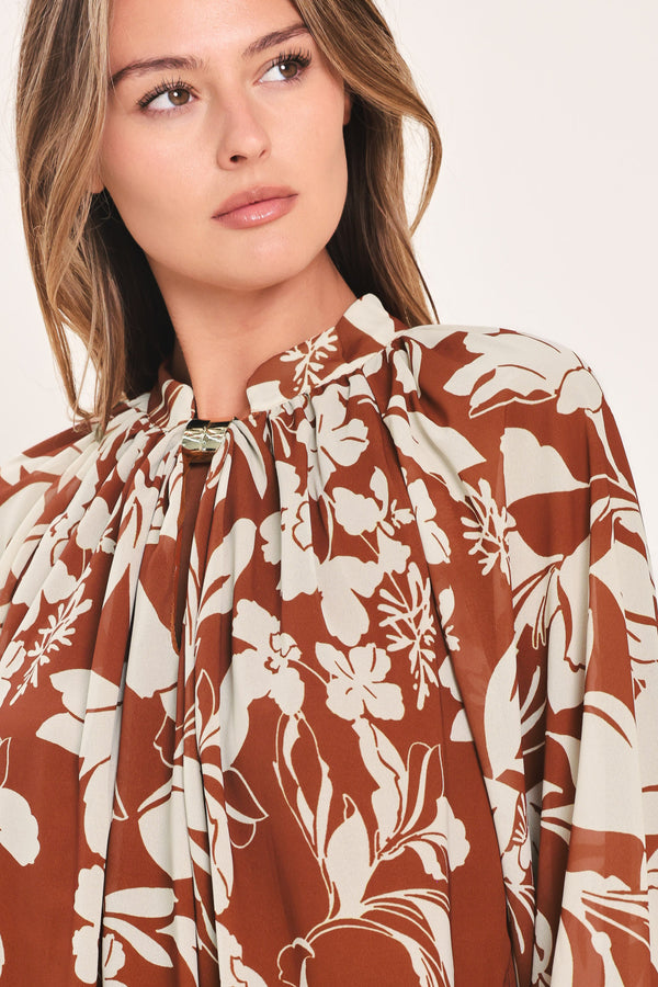 A woman wearing a rust-brown and cream floral print blouse with long sheer sleeves and a tie-neck detail | Flying Tomato