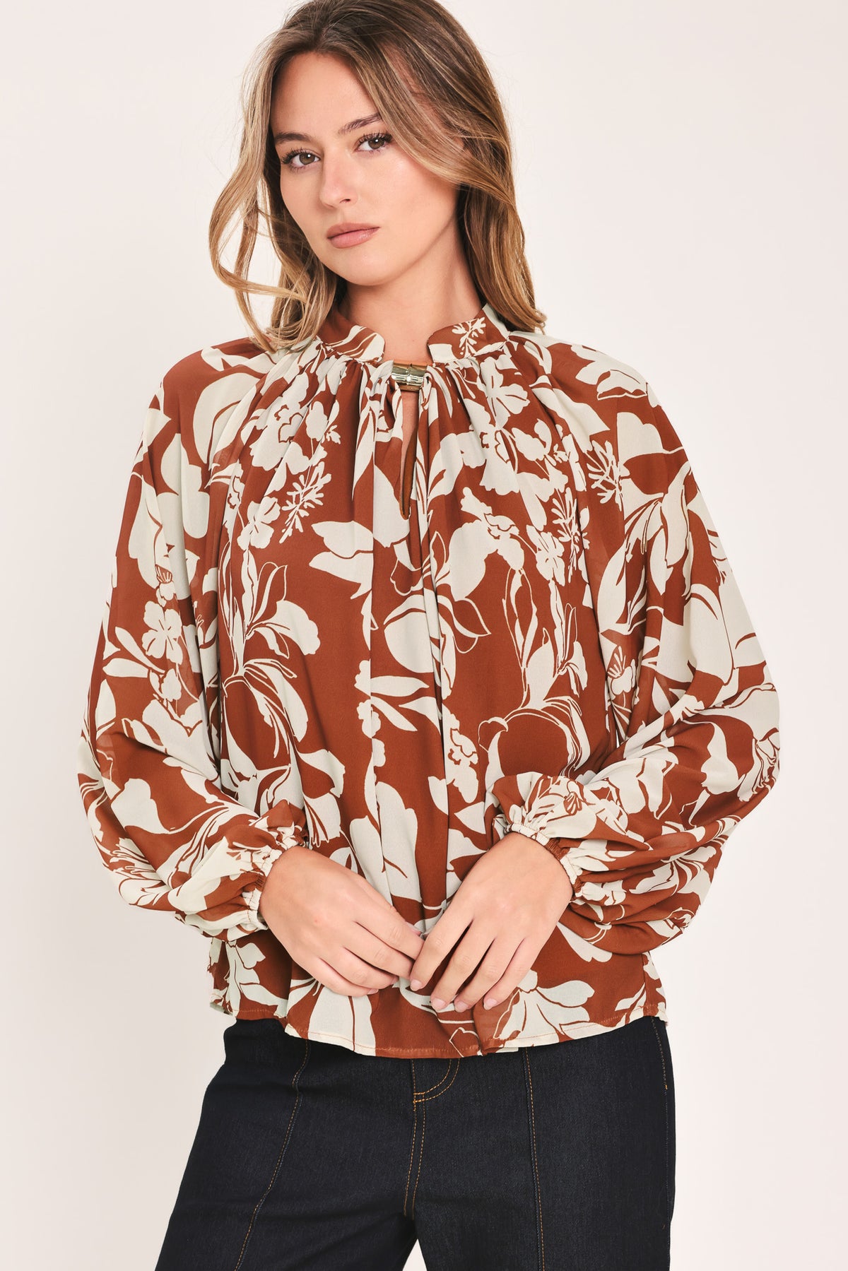 A woman wearing a rust-brown and cream floral print blouse with long sheer sleeves and a tie-neck detail | Flying Tomato