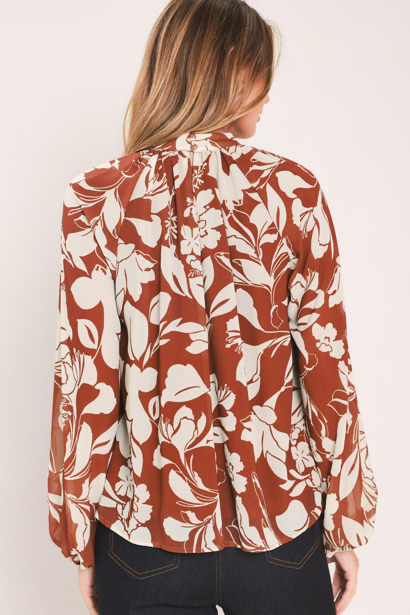 A woman wearing a rust-brown and cream floral print blouse with long sheer sleeves and a tie-neck detail | Flying Tomato