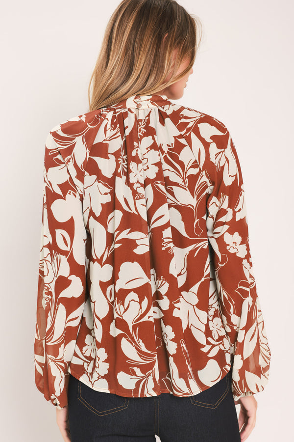 A woman wearing a rust-brown and cream floral print blouse with long sheer sleeves and a tie-neck detail | Flying Tomato