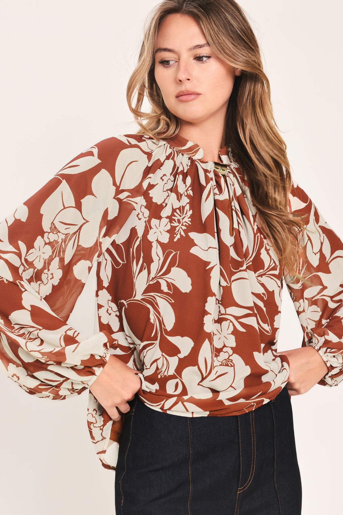 A woman wearing a rust-brown and cream floral print blouse with long sheer sleeves and a tie-neck detail | Flying Tomato