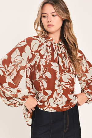 A woman wearing a rust-brown and cream floral print blouse with long sheer sleeves and a tie-neck detail | Flying Tomato