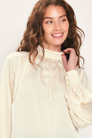 A solid woven top featuring a refined high neckline, long sleeves with buttoned cuffs, and a back button closure. 
