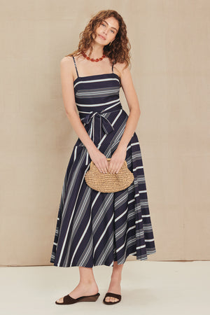 A woman modeling a navy blue midi dress with white and gray diagonal stripes, featuring thin spaghetti straps, a self-tie waist, and a flared skirt. She is holding a woven straw clutch and wearing dark brown slide sandals against a neutral beige background.