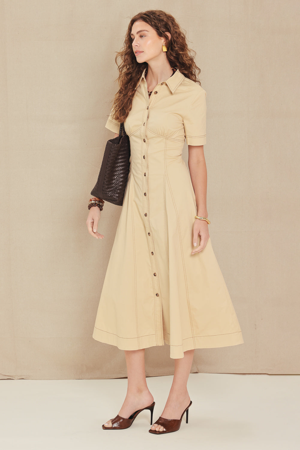 A woman models a short-sleeve midi shirt dress in a light beige color, styled with a black textured shoulder bag and brown heeled sandals against a neutral beige studio backdrop.