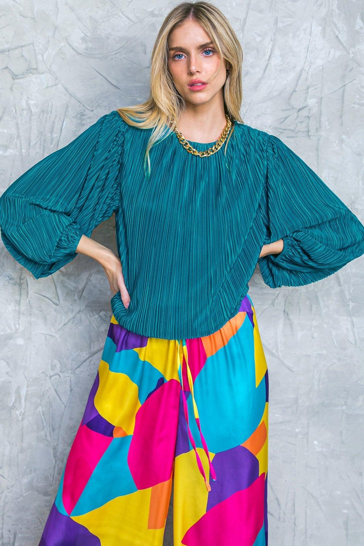 Woman wearing a bright pink pleated top and denim shorts sitting on a pink stool against a gray wall. FLYING TOMATO