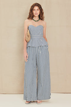 A woman models a strapless striped jumpsuit with a sweetheart bodice, peplum waist, and wide-leg pants in a blue and white vertical stripe pattern, standing against a neutral beige backdrop.