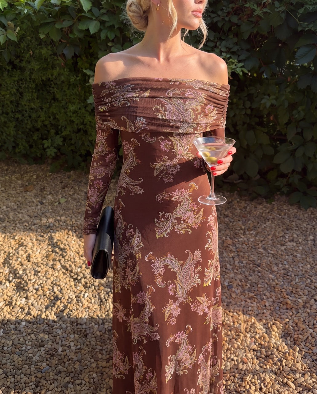 A woman outdoors wearing an off-the-shoulder long-sleeve maxi dress with a brown and beige baroque pattern. She is holding a black clutch and a martini glass, standing on a gravel path with green foliage in the background.